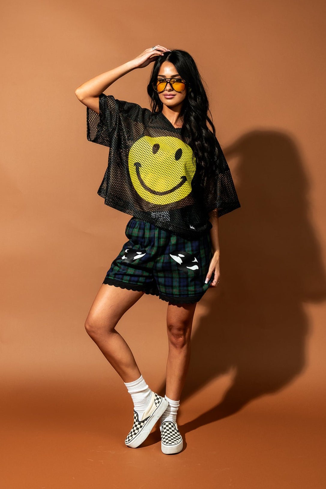 Girl/model standing against a brown backdrop wearing blue and green flannel boxer shorts with an orca graphic printed on each thigh with a black football jersey with a large yellow smile face printed on it
