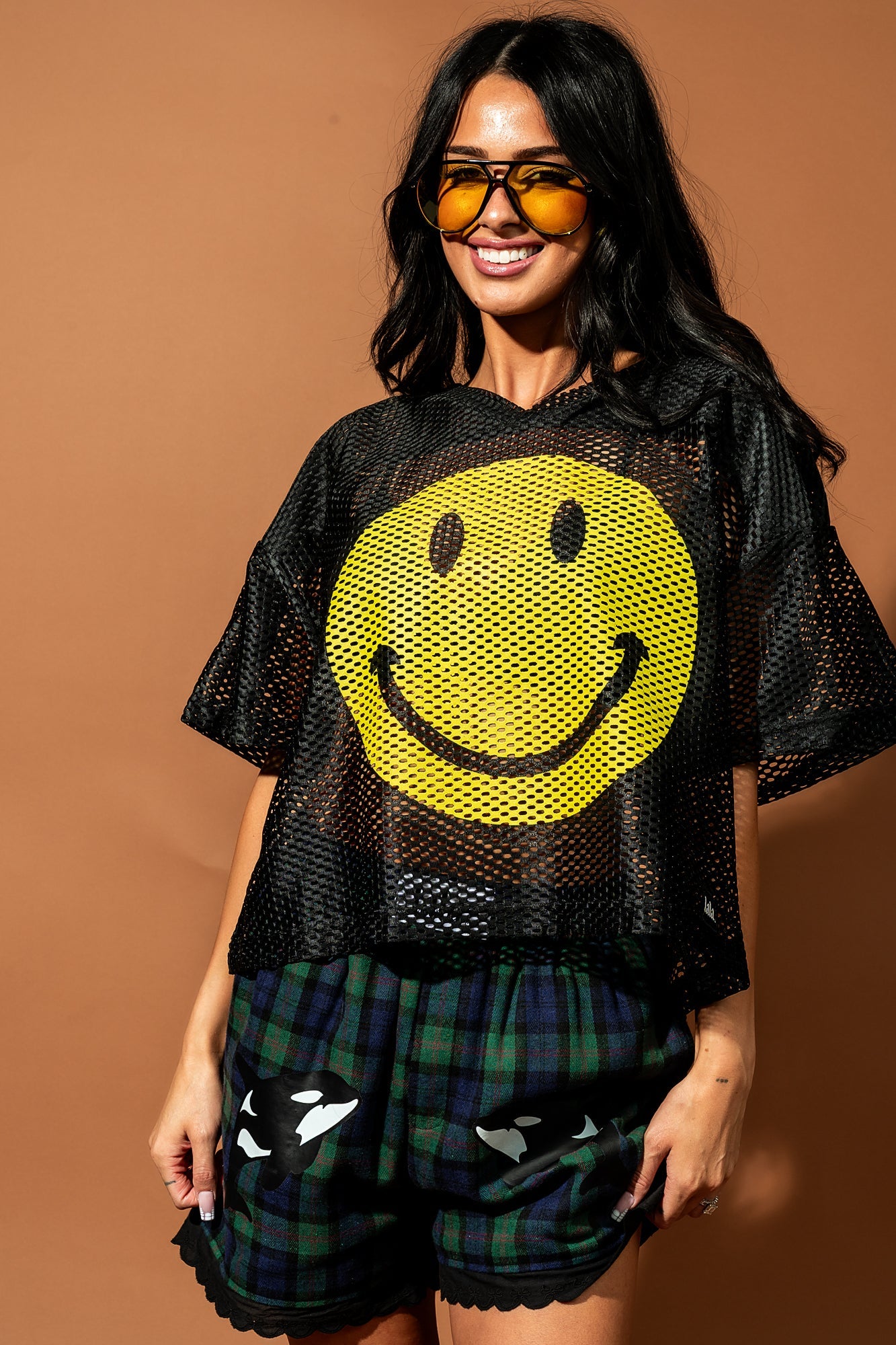 Girl/model standing against a brown backdrop wearing blue and green flannel boxer shorts with an orca graphic printed on each thigh with a black football jersey with a big smile face printed on it