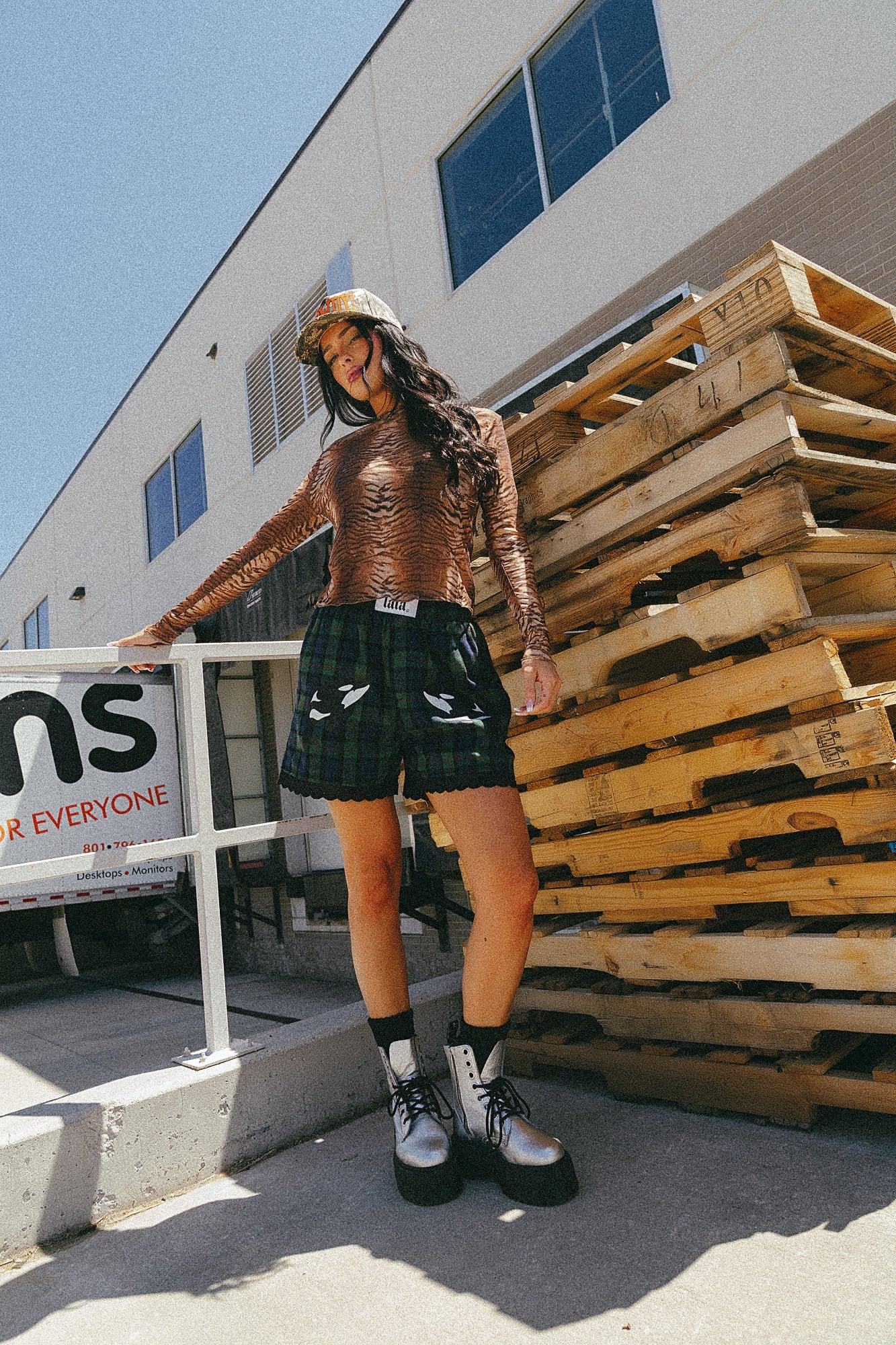 Girl/model standing outside against a stack of wooden pallets wearing blue and green flannel boxer shorts with an orca graphic printed on each thigh with tiger striped mesh top and a camo trucker hat
