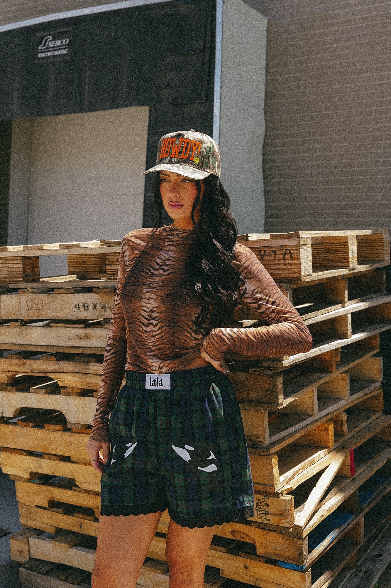 Girl/model standing outside against a stack of wooden pallets wearing blue and green flannel boxer shorts with an orca graphic printed on each thigh with tiger striped mesh top and a camo trucker hat