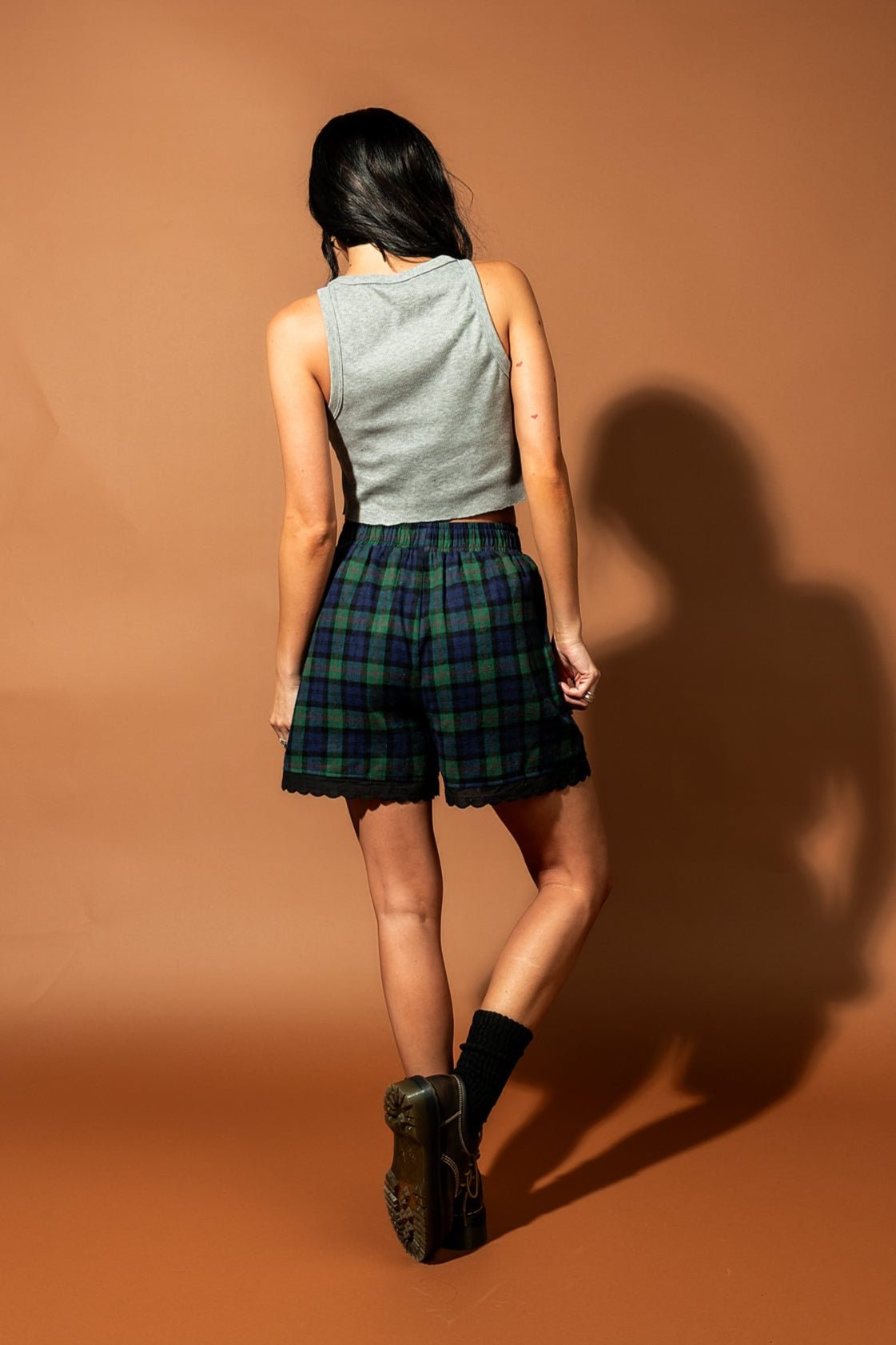 Girl/model standing against a brown backdrop wearing blue and green flannel boxer shorts with an orca graphic printed on each thigh with a grey tank top, facing away