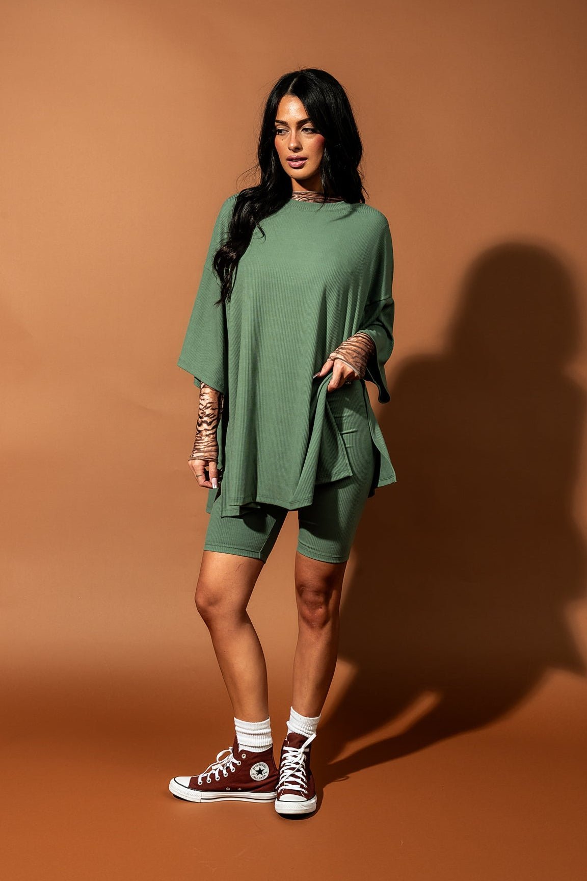 Girl/model standing against a brown backdrop wearing a sage green biker set with a tiger striped mesh top underneath