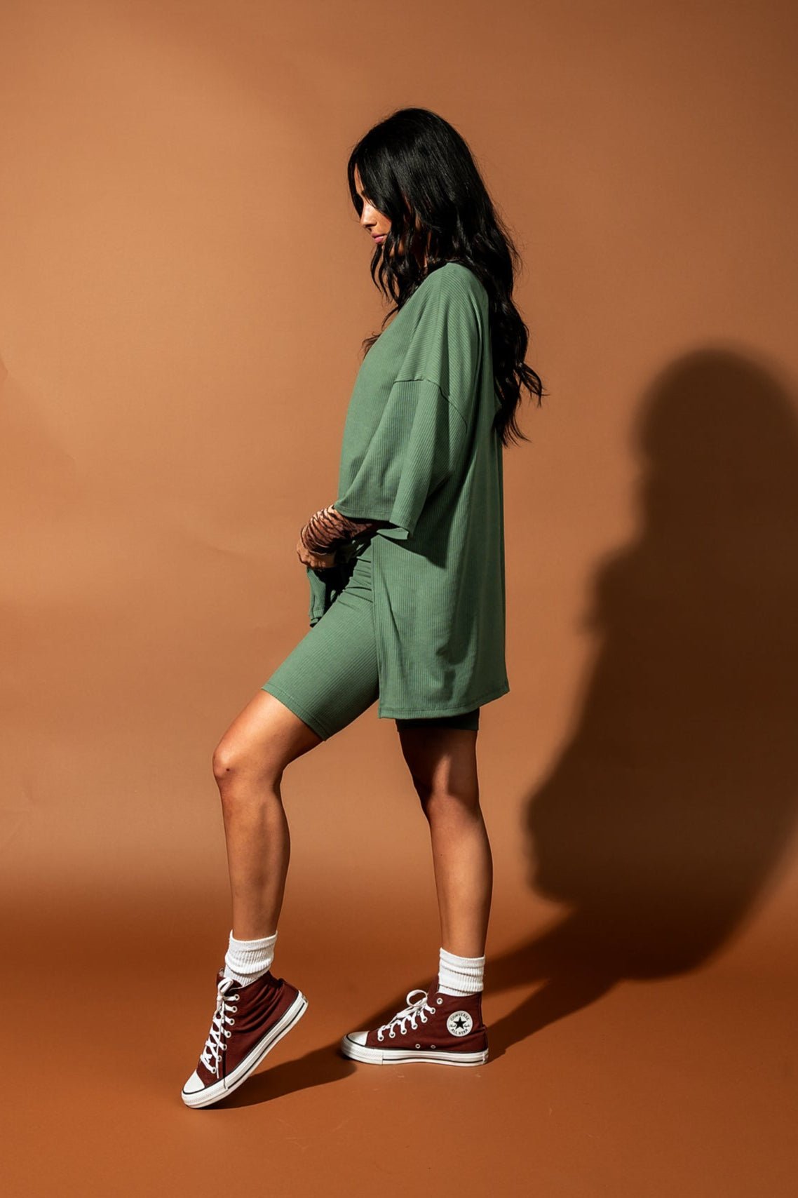 Girl/model standing against a brown backdrop wearing a sage green biker set with a tiger striped mesh top underneath