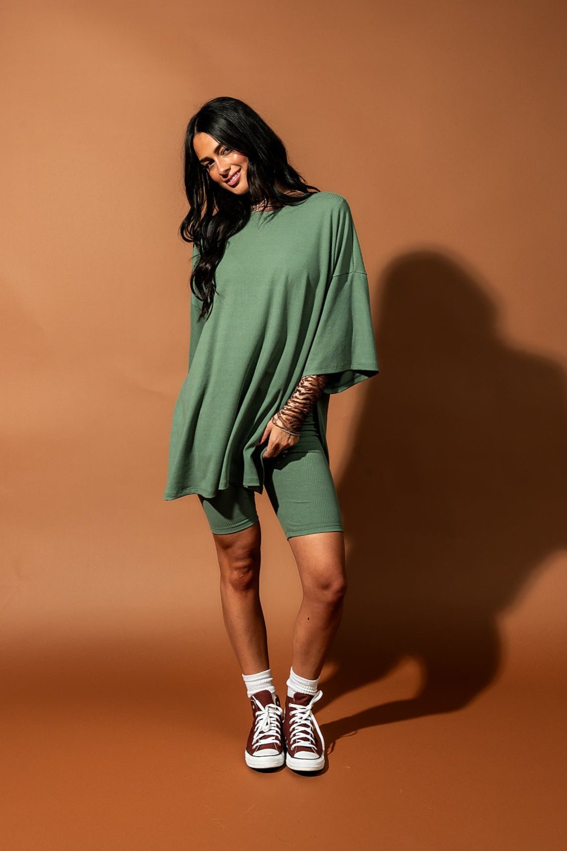 Girl/model standing against a brown backdrop wearing a sage green biker set with a tiger striped mesh top underneath