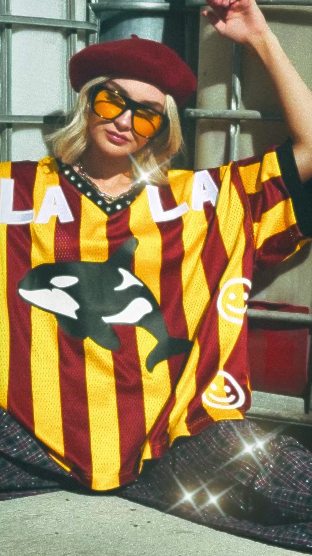 Totally Killer Orca Embellished Jersey - video of girl wearing rust + gold stripe jersey with orca, "LALA", and smiley faces on the front sitting on the ground outside leaning against old water containers