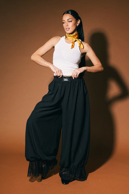 Woman wearing a white sleeveless top and black bloomer pants with a yellow scarf against a brown background