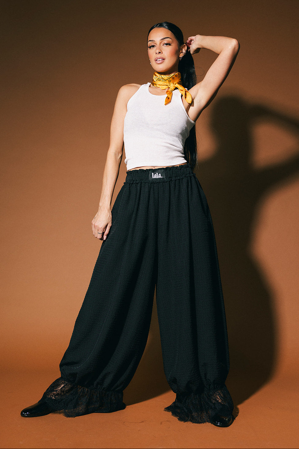 Woman wearing a white tank top and black bloomer pants with a yellow scarf on a brown background