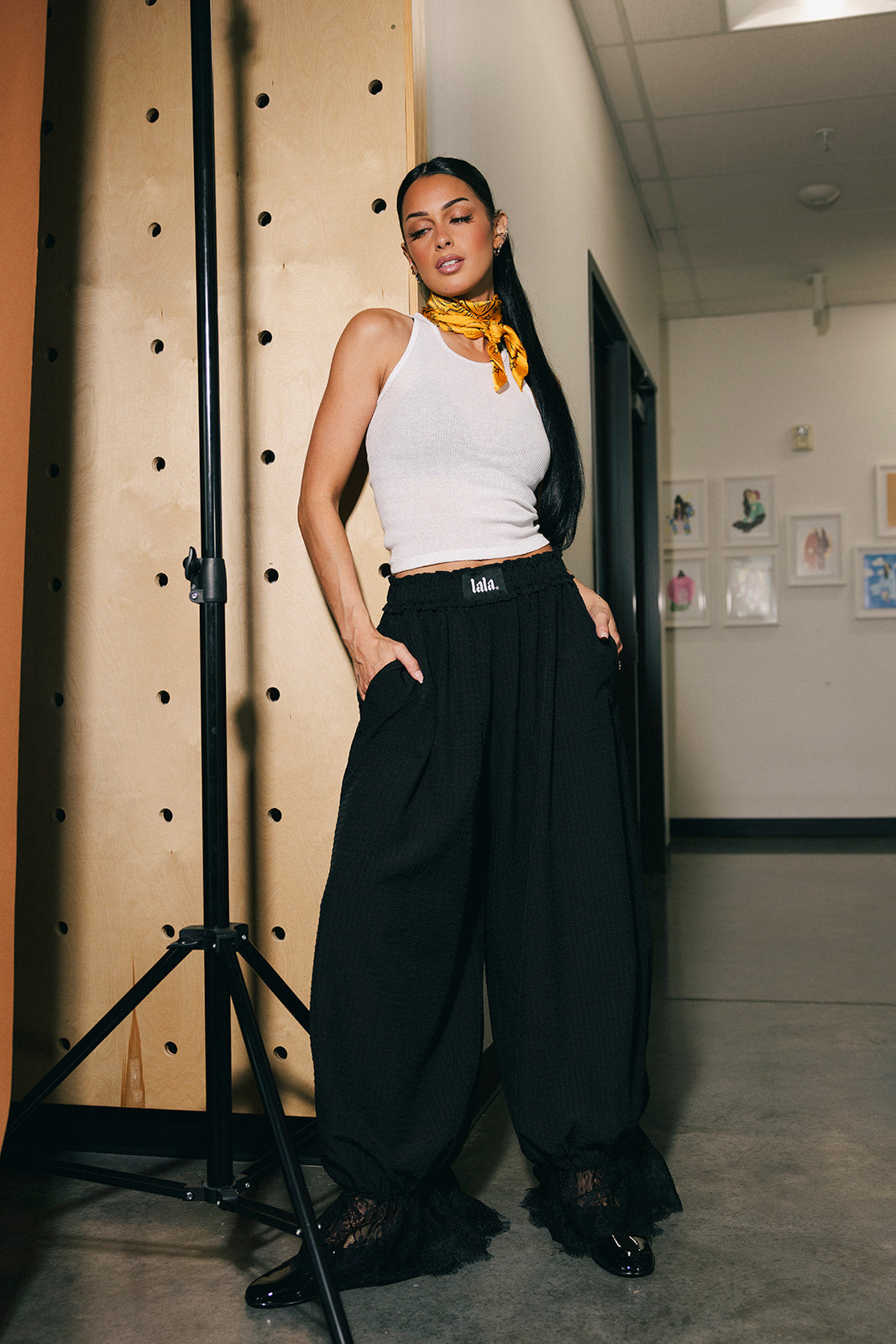 Woman in a white tank top and black bloomer pants standing in a room with a backdrop.