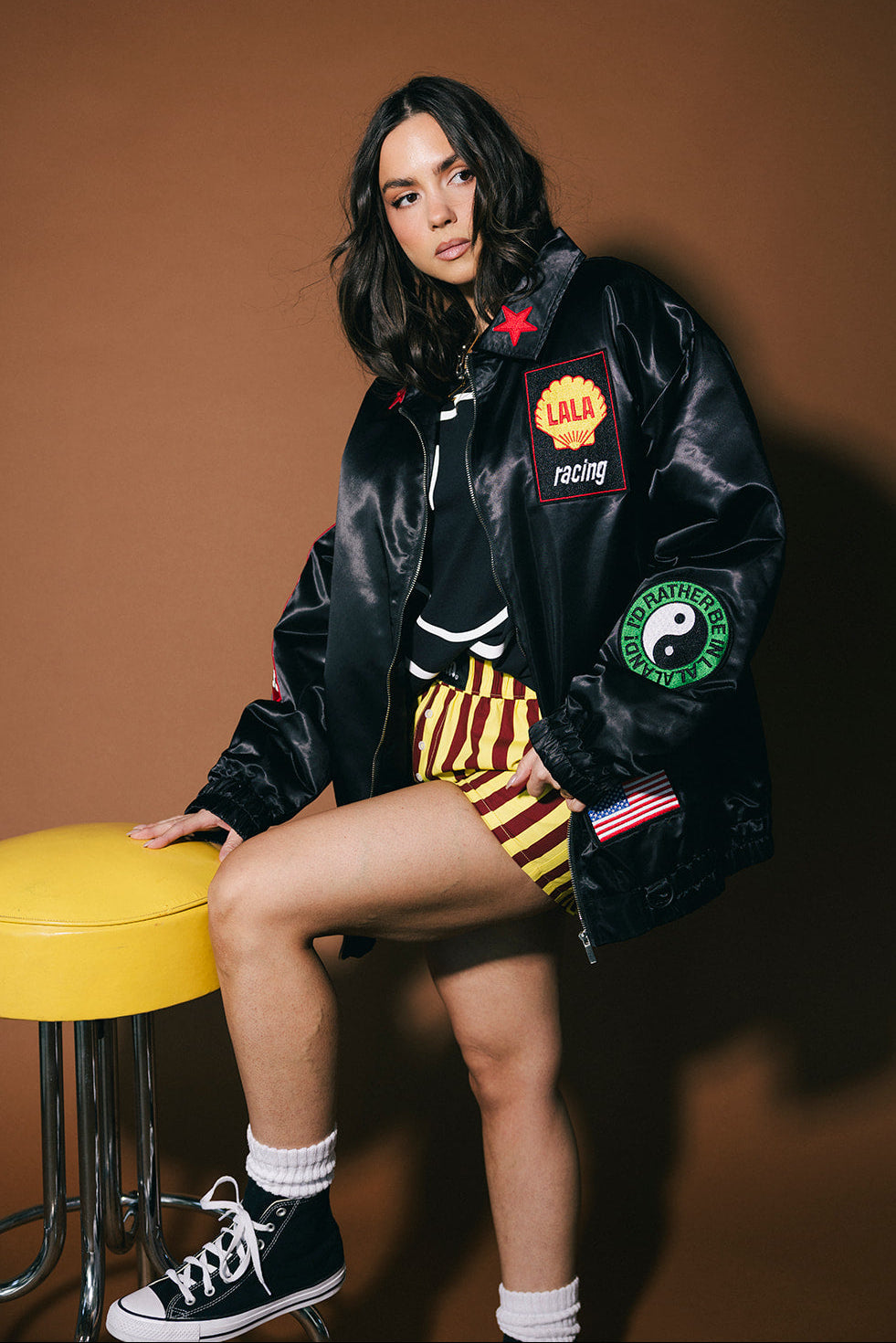 Person wearing a black jacket with patches, sitting on a yellow stool against a brown background