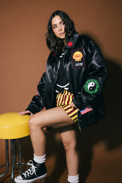 Person wearing a black jacket with patches, sitting on a yellow stool against a brown background