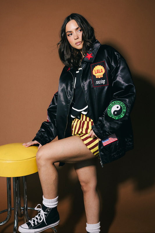Person wearing a black jacket with patches, sitting on a yellow stool against a brown background