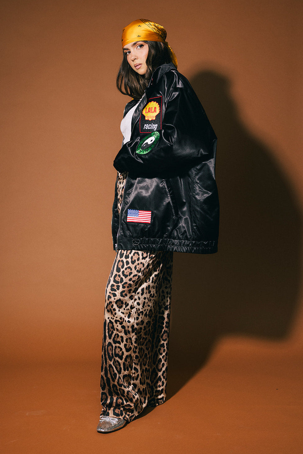 Person wearing a black jacket with patches, leopard print pants, and a yellow hat on a brown background