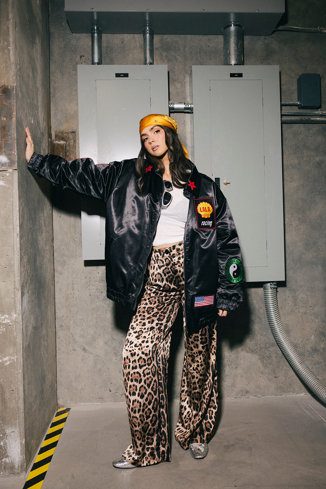 Person wearing a black jacket with patches, white shirt, and leopard print pants standing in an industrial setting.