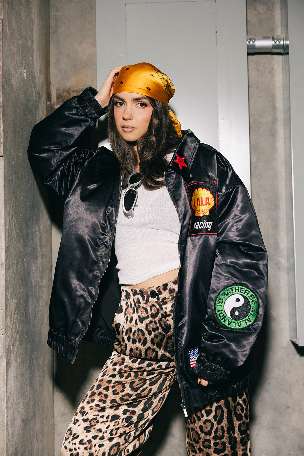 Person wearing a black jacket with patches, white top, and leopard print pants against a gray wall.