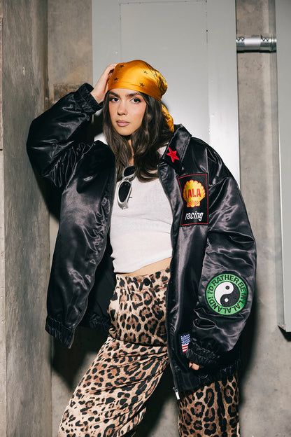 Person wearing a black jacket with patches, white top, and leopard print pants against a gray wall.