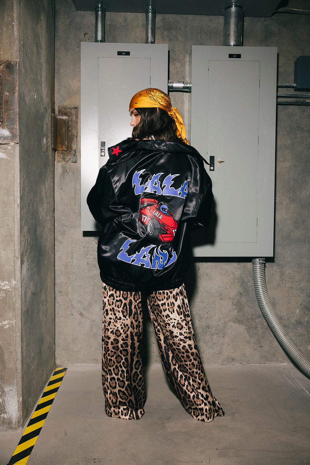 Person wearing a black jacket with colorful text and leopard print pants in an industrial setting
