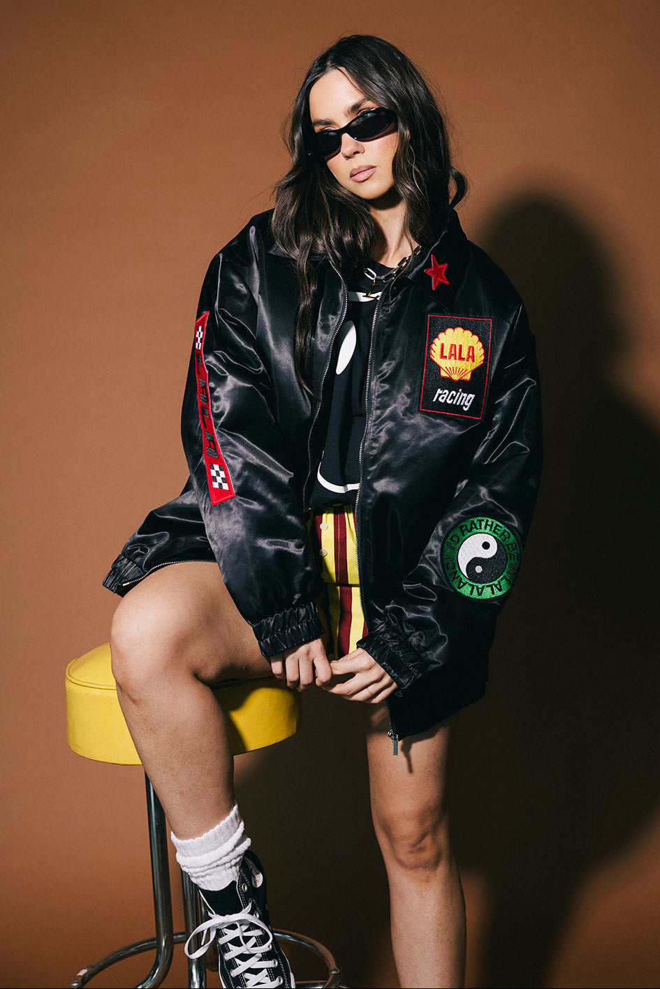 Person wearing a black jacket with patches sitting on a yellow stool against a brown background