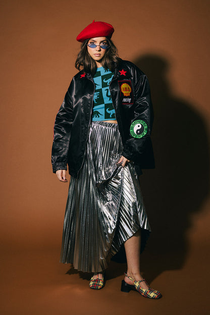 Person wearing a black jacket with colorful patches, silver pleated skirt, and red beret on a brown background