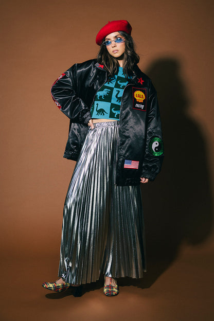 Person wearing a black jacket with patches, blue top, silver pleated skirt, and red beret against a brown background