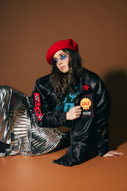 Person wearing a black jacket with patches, silver pants, and a red beret on a brown background