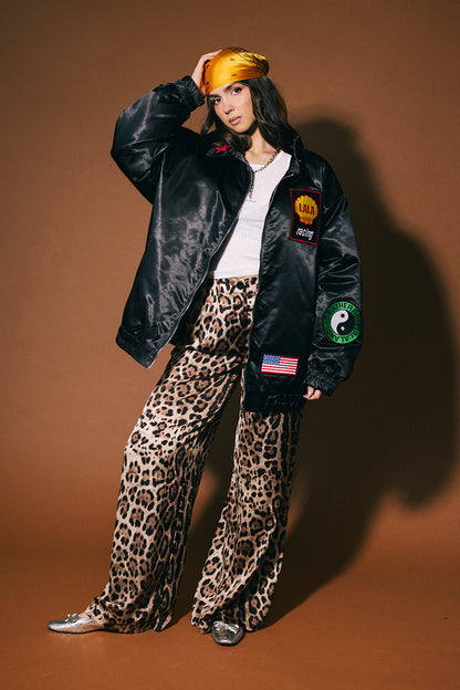 Person wearing a black jacket with patches and leopard print pants on a brown background