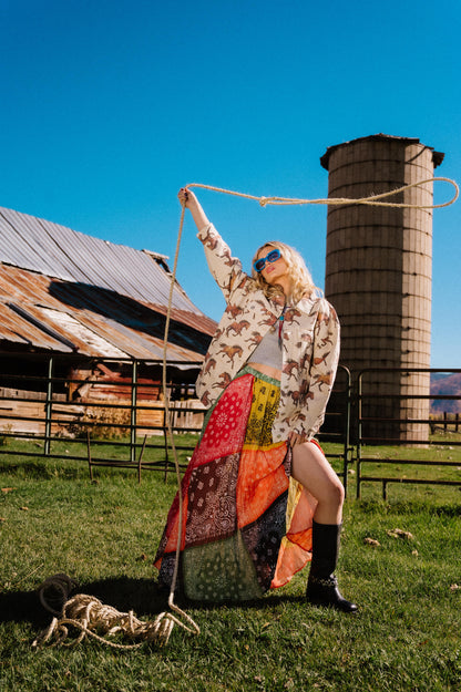 Person holding a lasso in front of a barn and silo on a sunny day wearing a patchwork style bandana print pleated midi skirt with a side slit