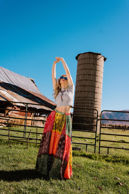 Person in patchwork style bandana print pleated midi skirt standing in a rural setting with a silo and barn in the background.