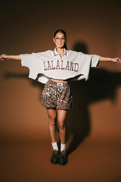 Person wearing a 'LALALAND' oversized polo and leopard print bloomer shorts with arms outstretched on a brown background