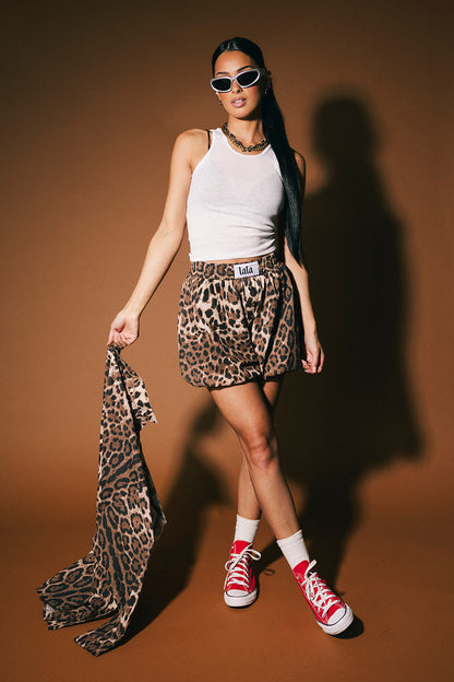 Woman in leopard print bloomer shorts and white top holding a matching top against a brown background