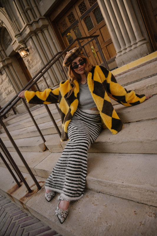 Woman in a yellow and brown diamond faux fur coat sitting on stone steps in front of a building.