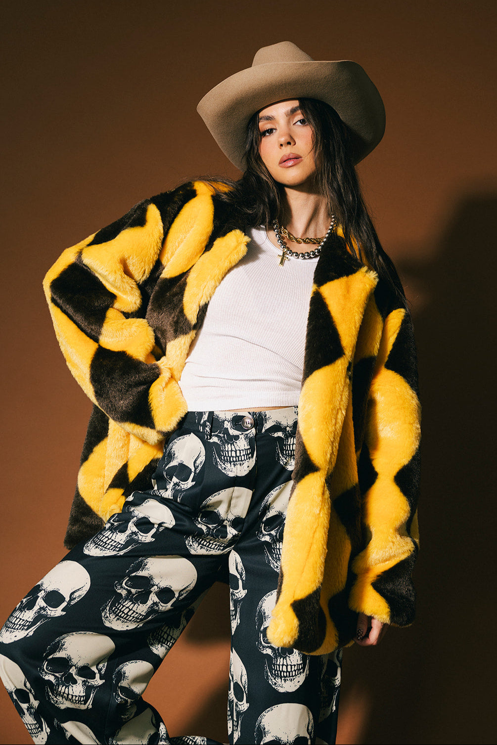 Person wearing a yellow and brown diamond faux fur coat with a white shirt and skull-patterned trousers on a brown background
