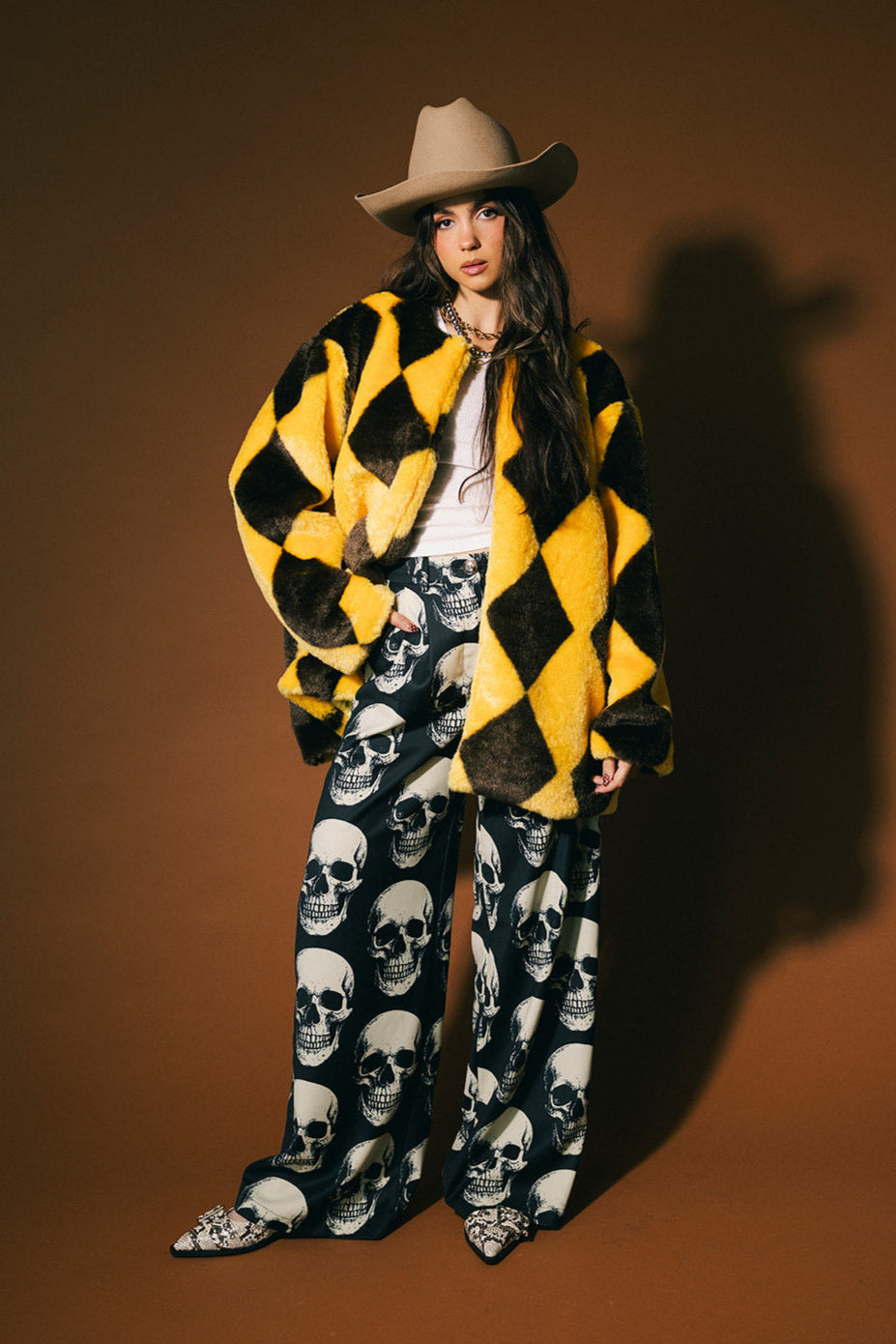 Person wearing a yellow and brown diamond faux fur coat, beige cowboy hat, and skull-patterned trousers on a brown background