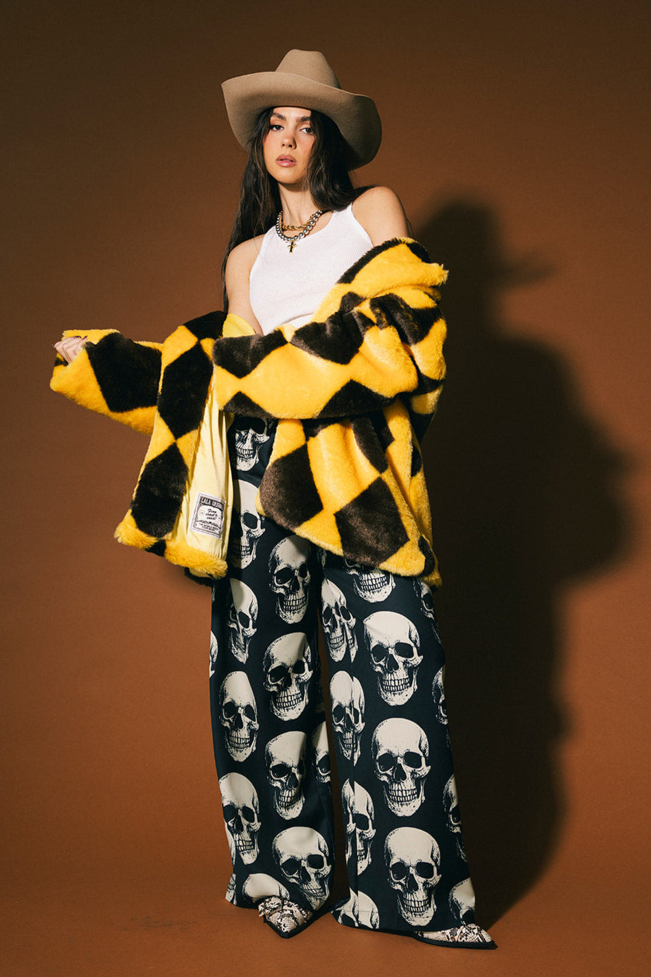 Woman wearing a yellow and brown diamond faux fur coat, white top, and skull-patterned trousers against a brown background