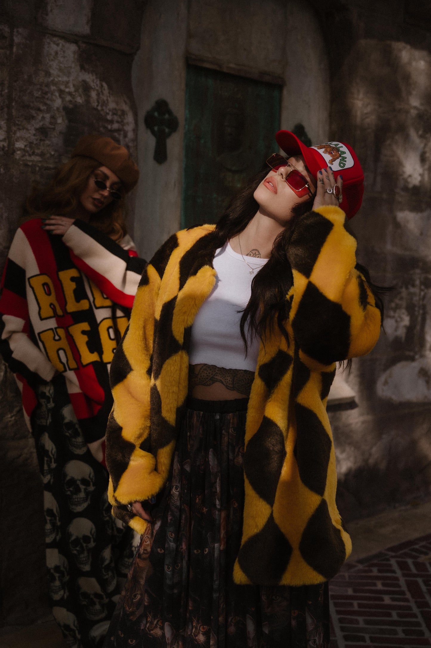 Two people in front of a building. One is wearing a yellow and brown diamond faux fur coat and red trucker hat.