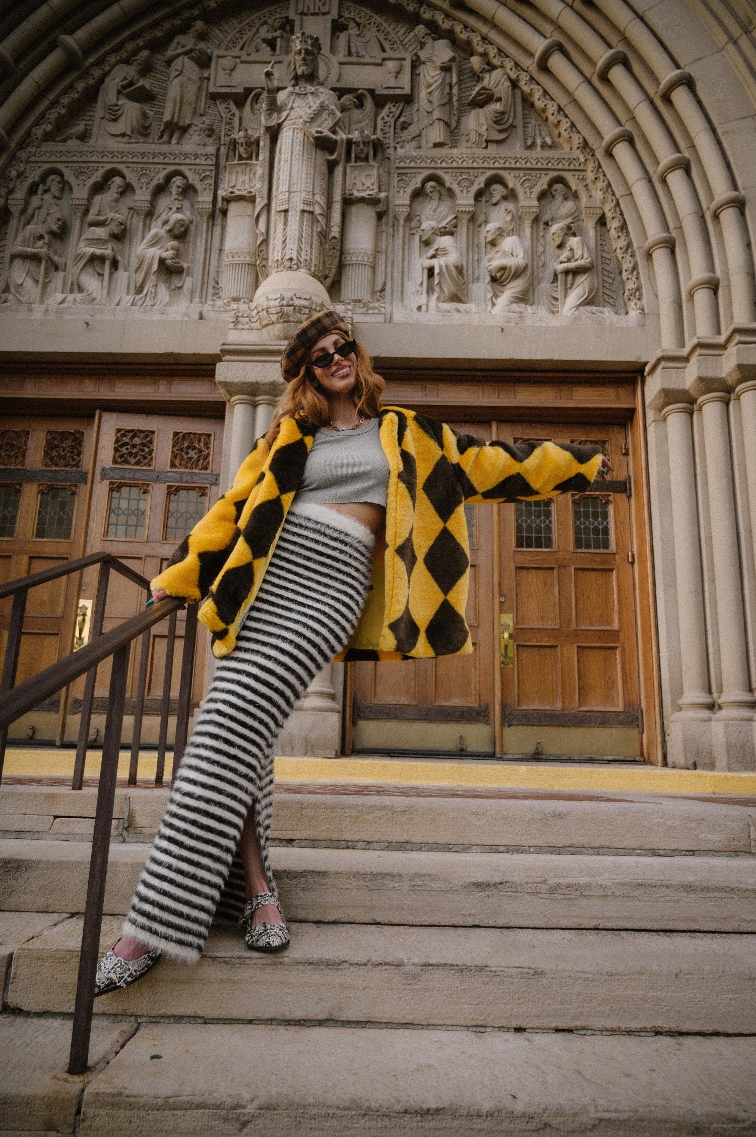 Person posing on steps in front of a decorative building entrance wearing a yellow and brown diamond faux fur coat.
