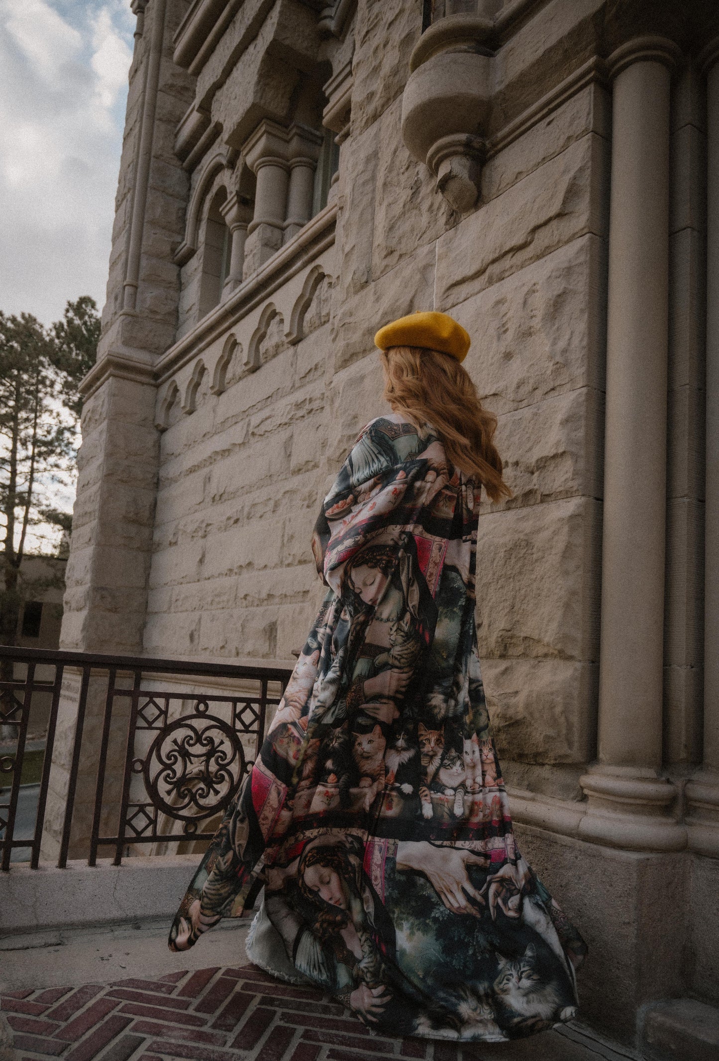 Woman in a renaissance cat pattern satin duster and yellow beret standing in front of a stone building.