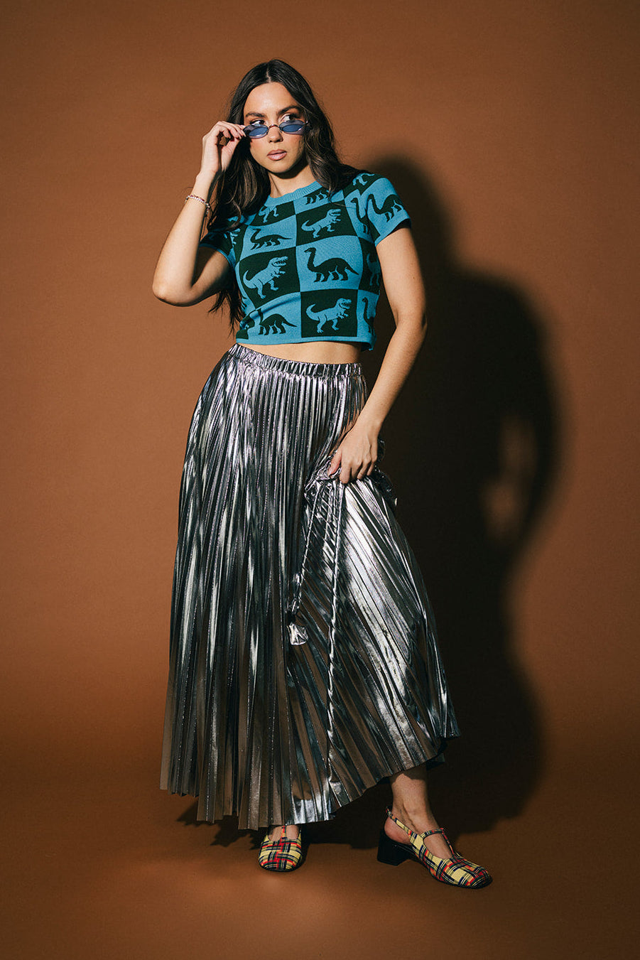 Woman wearing a blue top with dinosaur patterns and a silver pleated skirt against a brown background