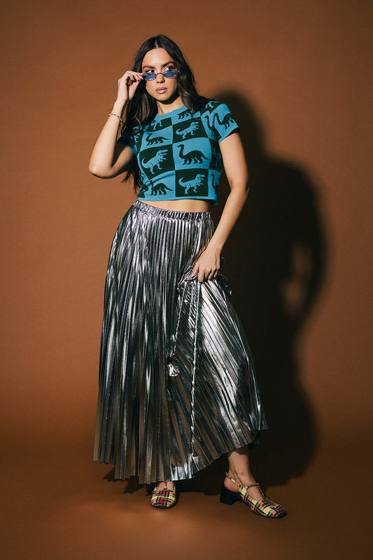 Woman wearing a blue top with dinosaur patterns and a silver pleated skirt against a brown background