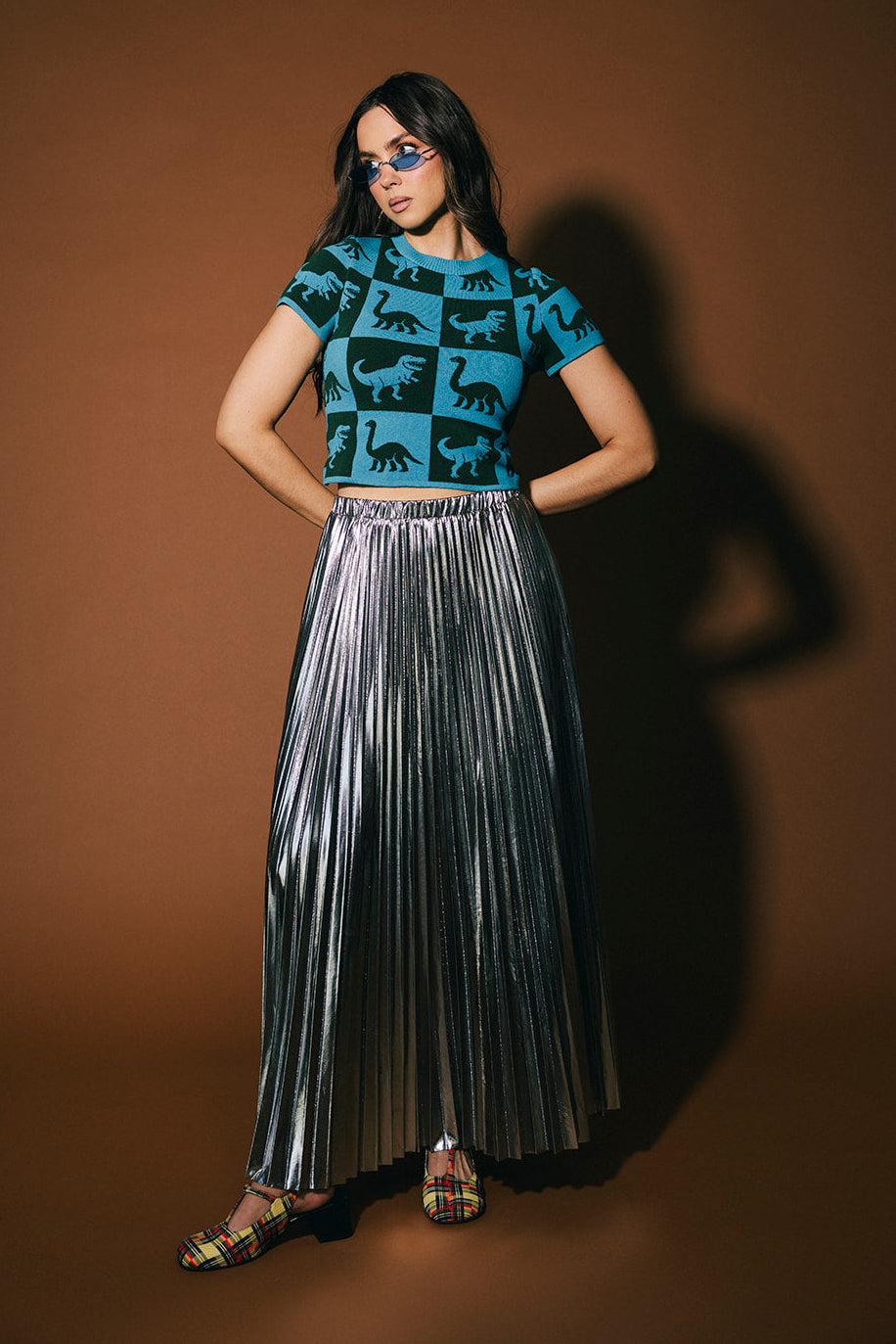 Woman wearing a blue patterned top and silver pleated skirt against a brown background