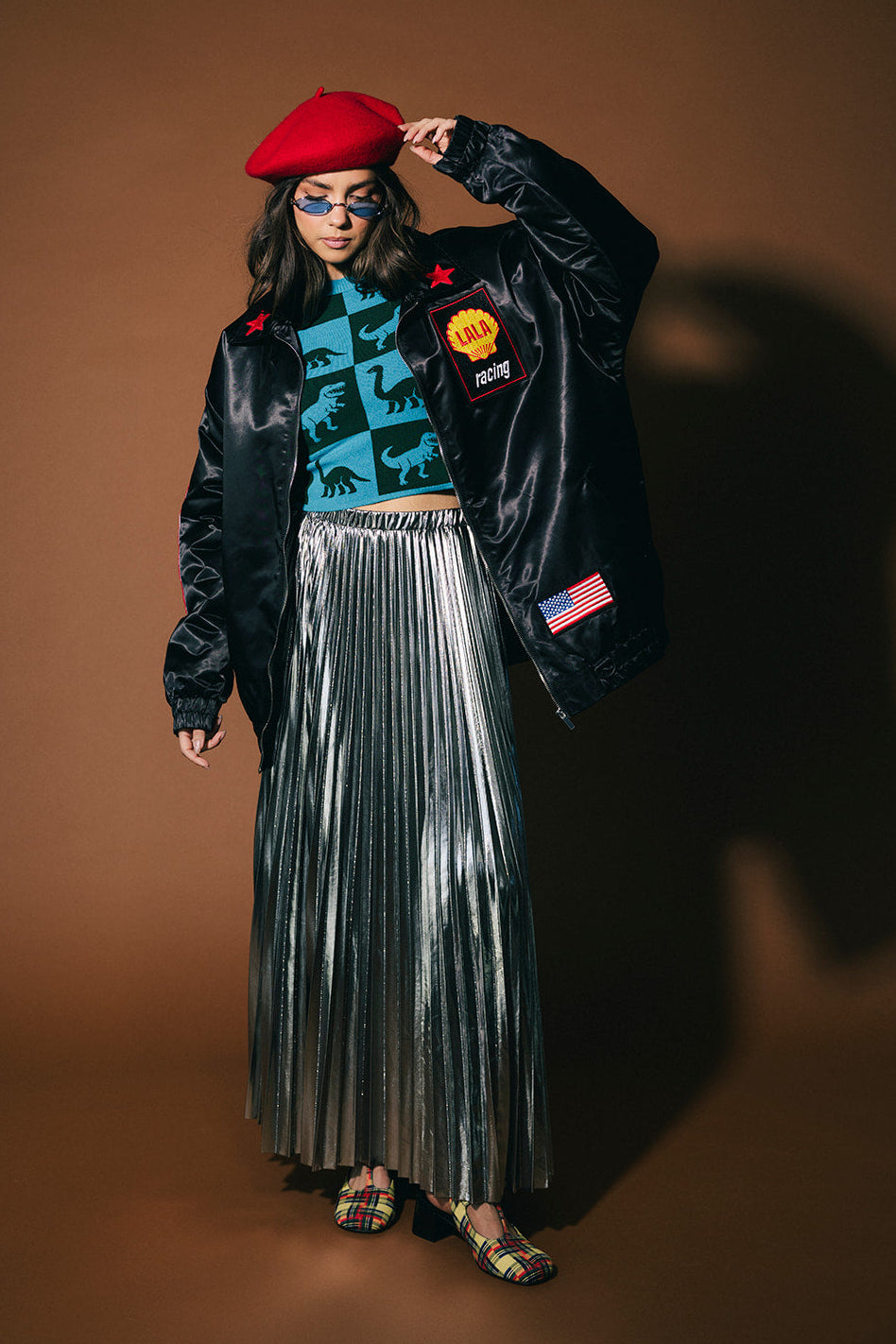 Person wearing a black jacket with patches, blue patterned shirt, red beret, and silver pleated skirt on a brown background