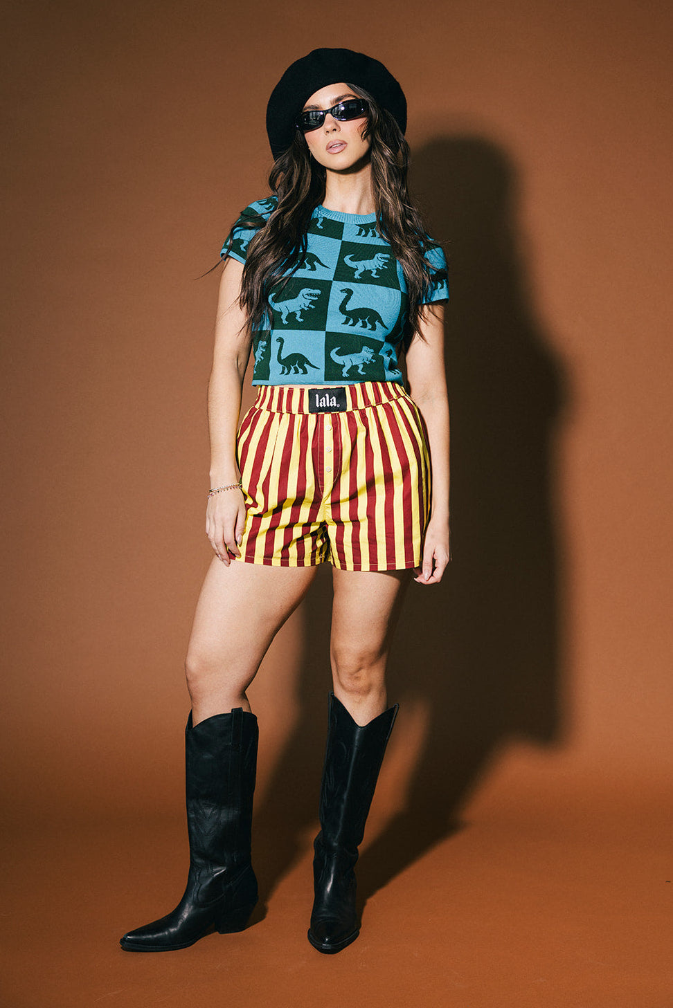 Woman wearing a dinosaur patterned top, striped shorts, and black boots against a brown background