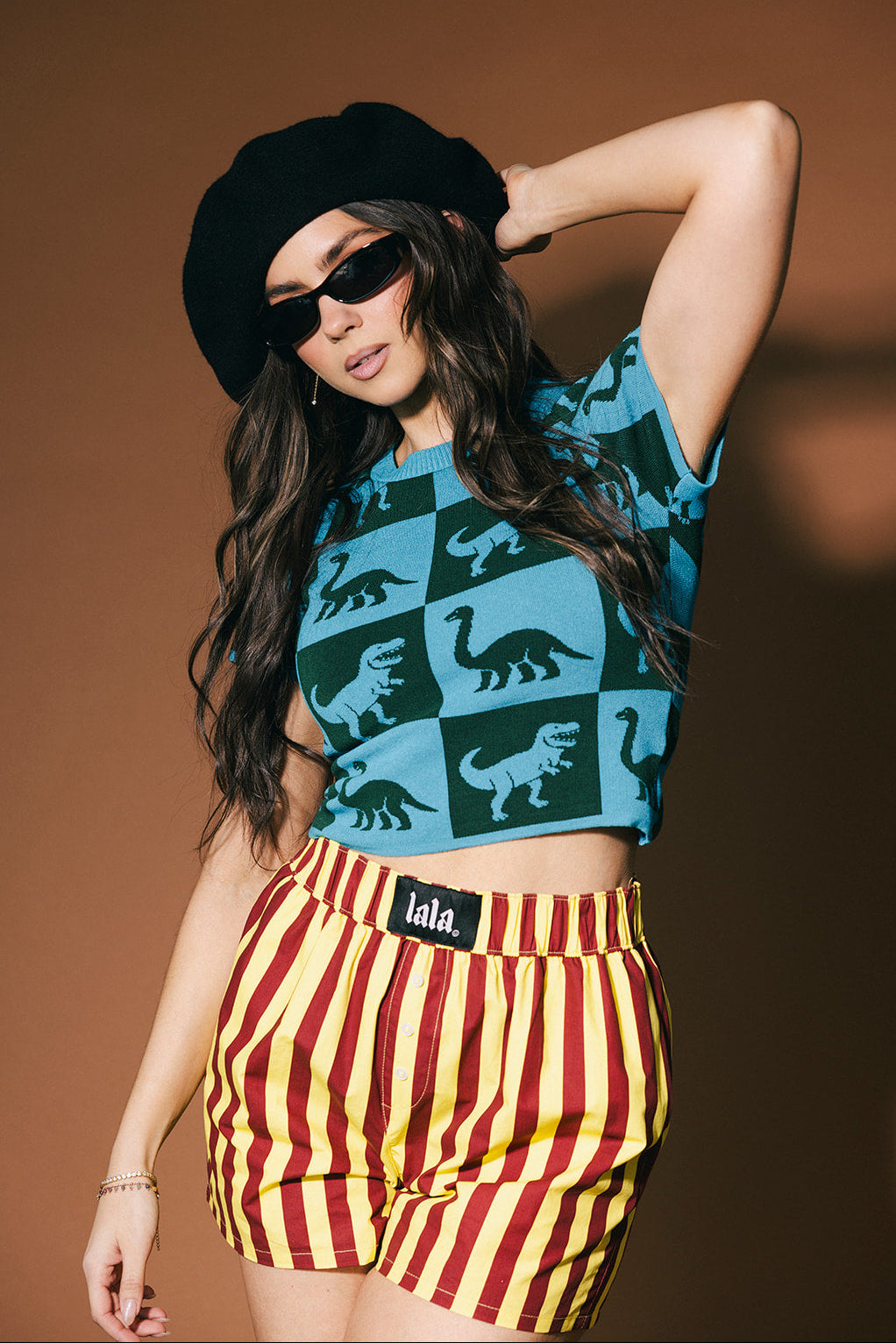 Woman wearing a blue checkered top with dinosaur patterns and red and yellow striped shorts on a brown background.