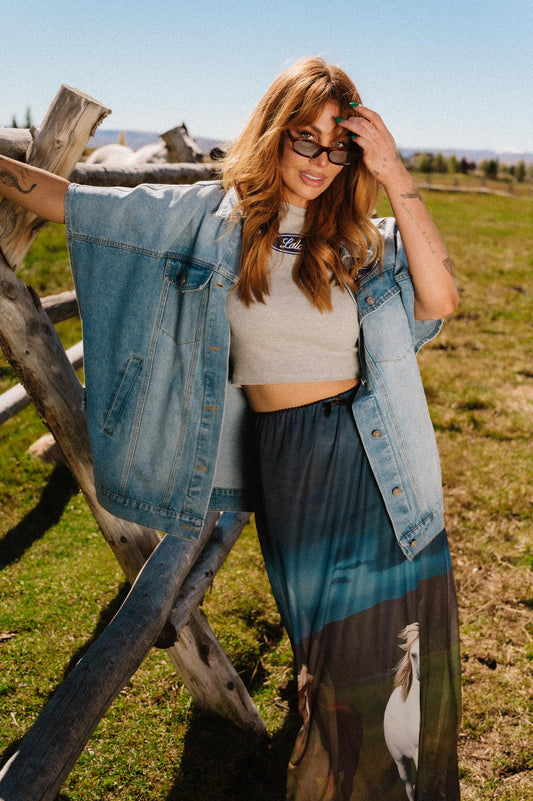 Woman standing outdoors near a wooden fence wearing sunglasses and a denim jacket.