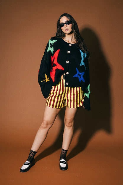 Woman wearing a colorful cardigan, striped shorts, and sunglasses on a brown background