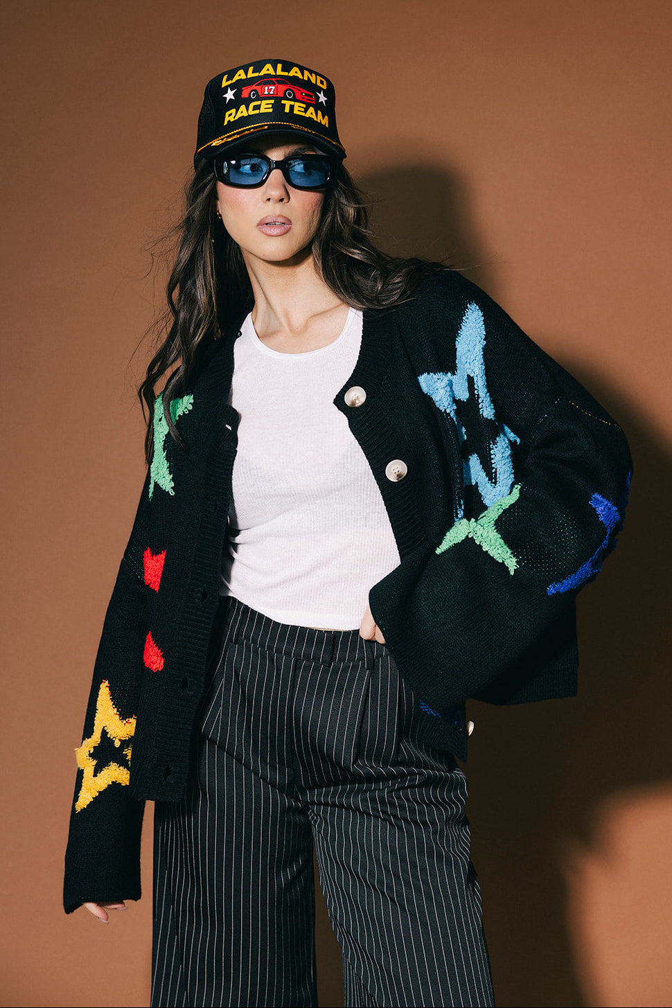 Person wearing a colorful cardigan with star patterns, white shirt, and black pants against a brown background.