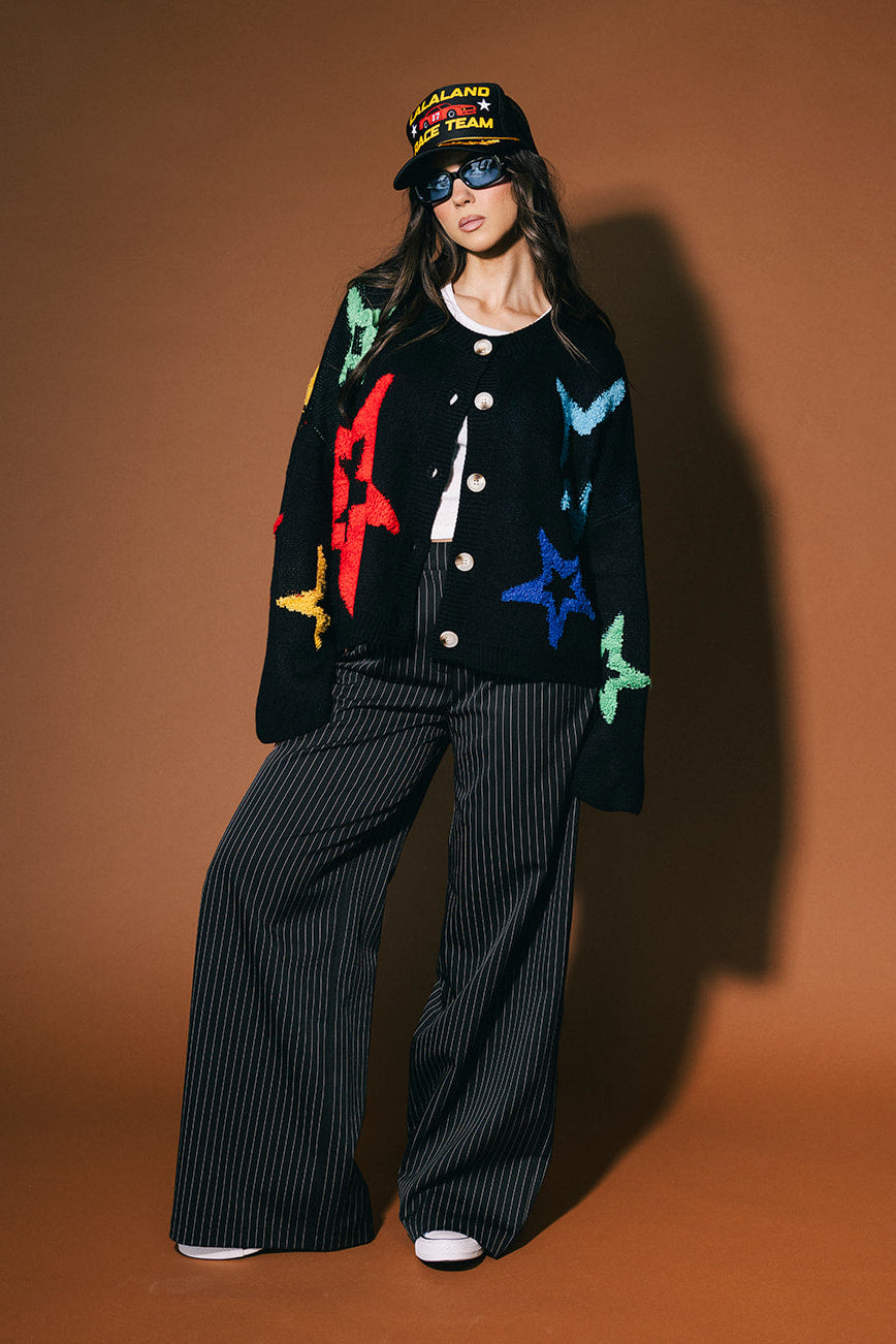 Person wearing a black cardigan with colorful star patterns, black pants, and a black cap on a brown background