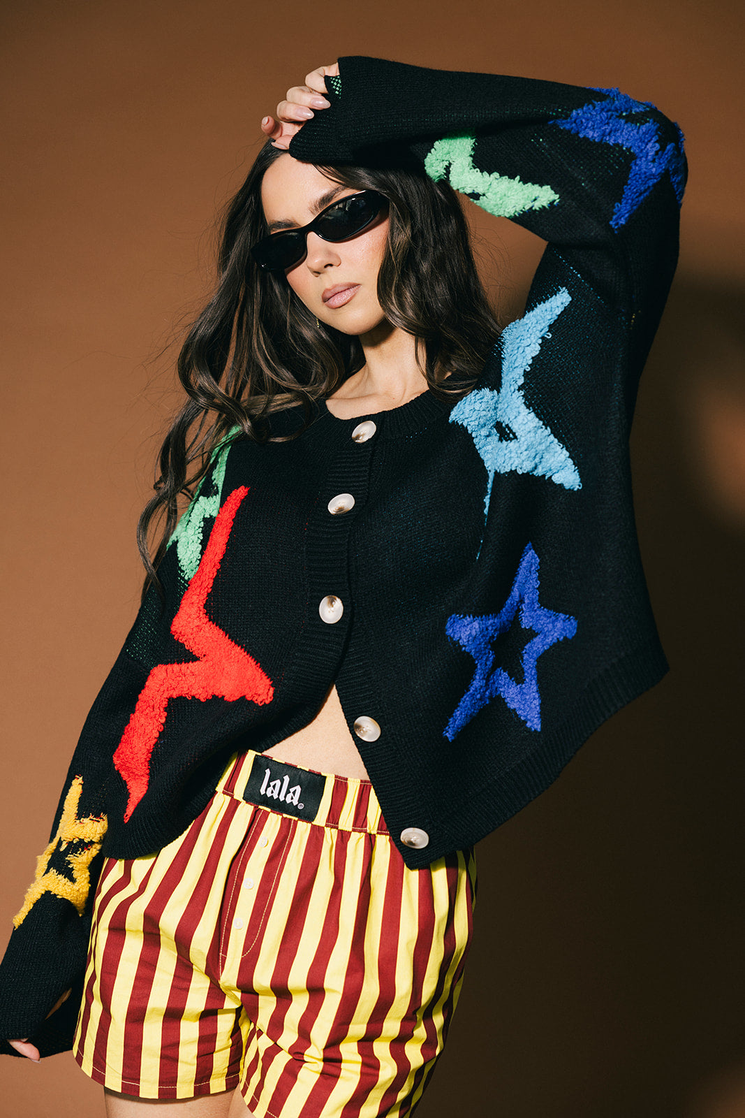 Person wearing a colorful cardigan with star patterns and striped shorts against a brown background