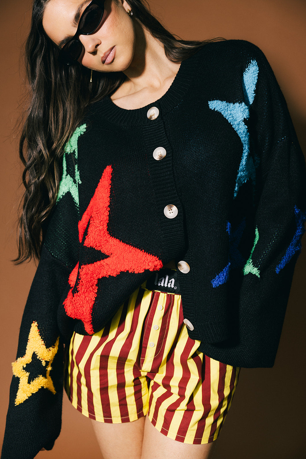 Person wearing a black cardigan with colorful star patterns and striped shorts against a brown background