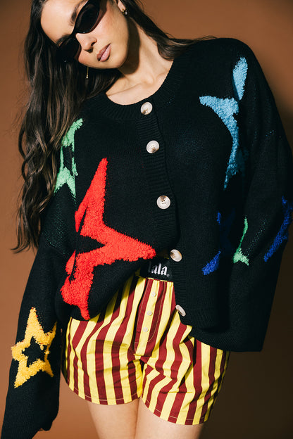 Person wearing a black cardigan with colorful star patterns and striped shorts against a brown background