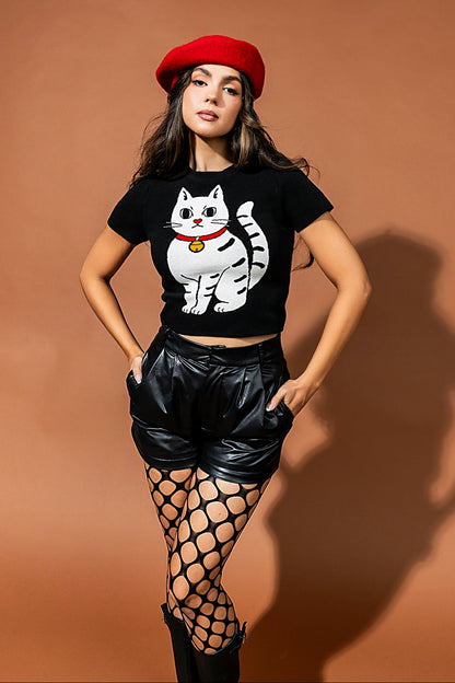 Woman wearing a black t-shirt with a cat graphic, red beret, black shorts, and fishnet stockings against a brown background.
